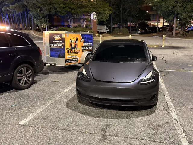 Gray Tesla sedan near the Trustee branded trailer in a parking lot at night