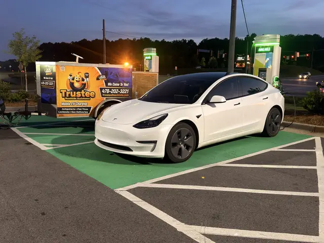 White Tesla parked near the Trustee trailer at a public charging site at dusk